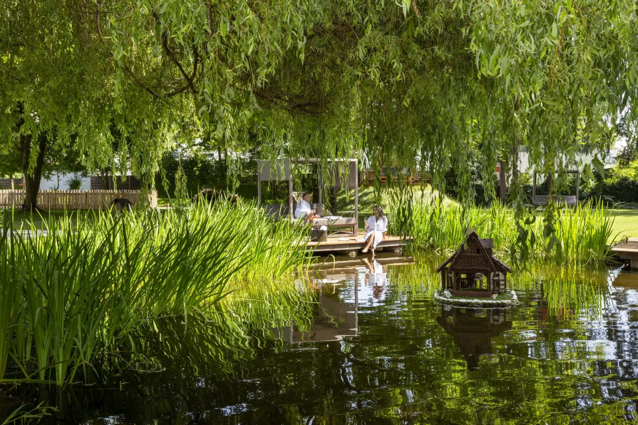 Entspannung am Teich: Gäste in Bademänteln auf Holzdeck unter Trauerweide im idyllischen Garten des Das Parkhotel.