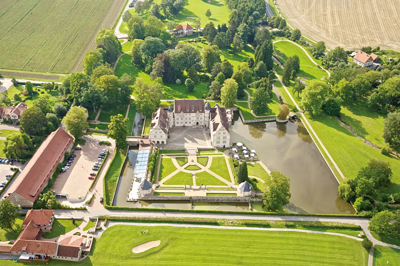 Malerische Luftaufnahme des historischen Schlosshotel Münchhausen mit Wassergraben, Barockgarten und weitläufiger Parklandschaft.