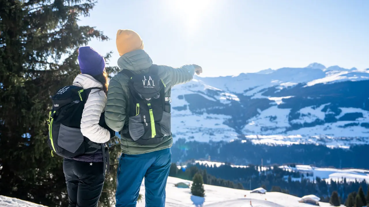 Zwei Wanderer mit Rucksäcken und La Val Hotel & Spa Logo blicken auf verschneite Schweizer Alpenlandschaft.