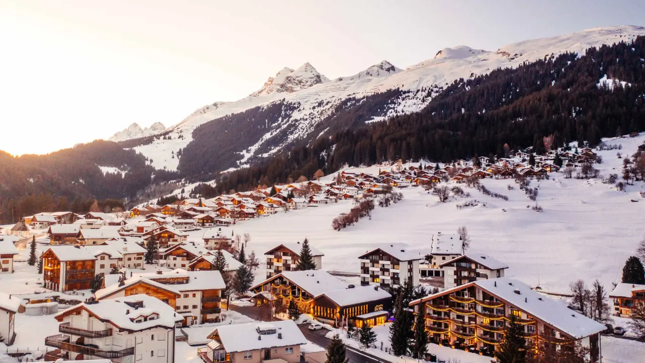 Winterpanorama des La Val Hotel & Spa in einem verschneiten Bergdorf mit beleuchteten Fassaden vor majestätischen Alpen.