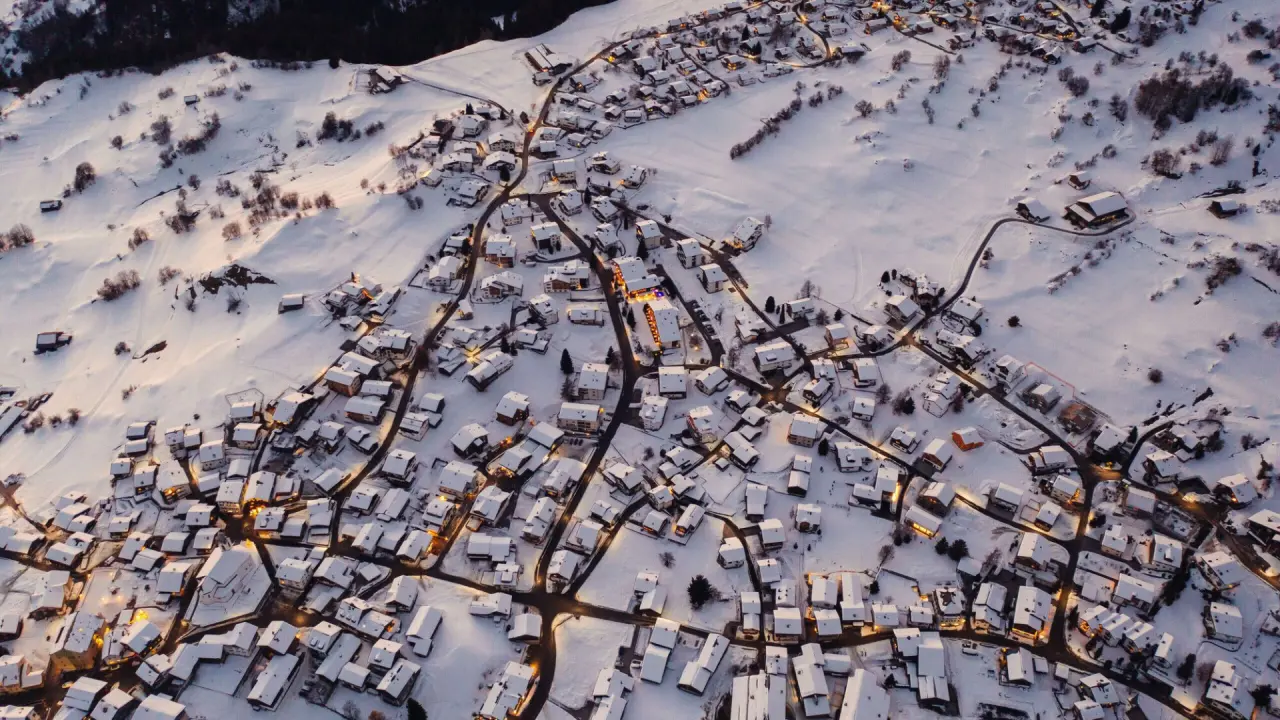 Luftaufnahme des verschneiten Bergdorfes mit beleuchteten Häusern bei Dämmerung, inklusive La Val Hotel & Spa.