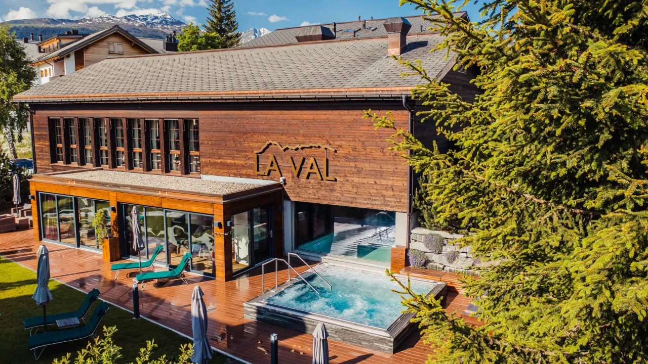 Aussenansicht des La Val Hotel & Spa mit Holzfassade, Aussen-Whirlpool, Sonnenliegen und schneebedeckten Bergen.