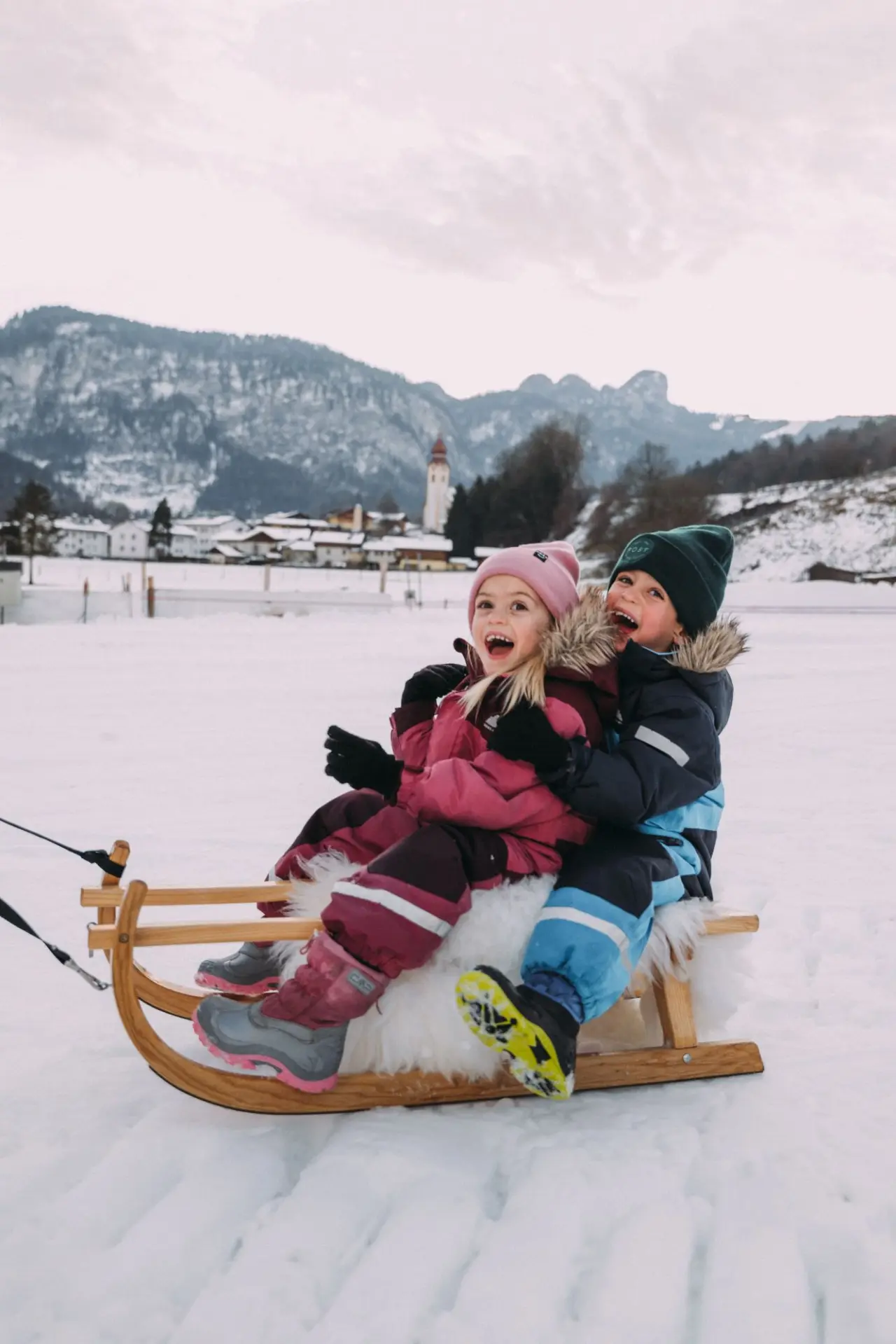 Zwei lachende Kinder auf einem Schlitten im Schnee vor Bergkulisse im POST Family Resort.
