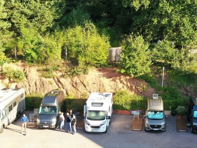 Parkplatz mit mehreren Wohnmobilen und Campern vor einem bewaldeten Hang am Hotel Am Hirschhorn.