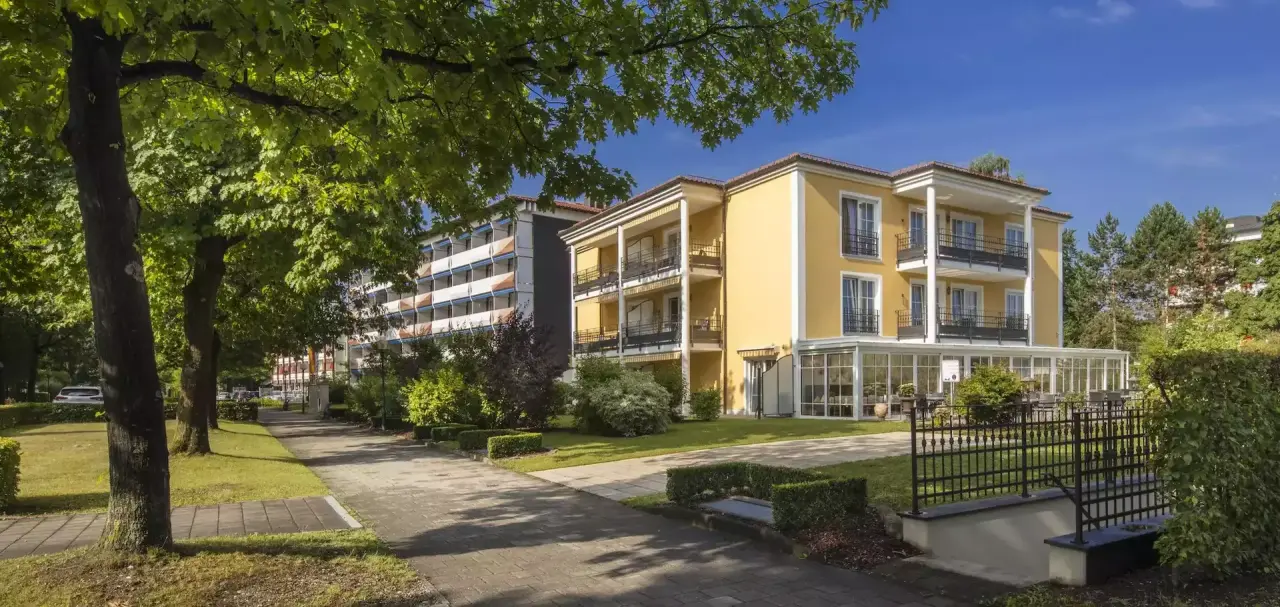 Aussenansicht des Hotel Schweizer Hof Betriebs-GmbH mit gelber Fassade, Balkonen und grünen Bäumen unter blauem Himmel.