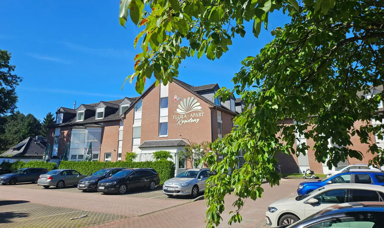 Aussenansicht der Flora-Apart Residenz mit roter Backsteinfassade, Parkplatz und grünen Bäumen an einem sonnigen Tag.