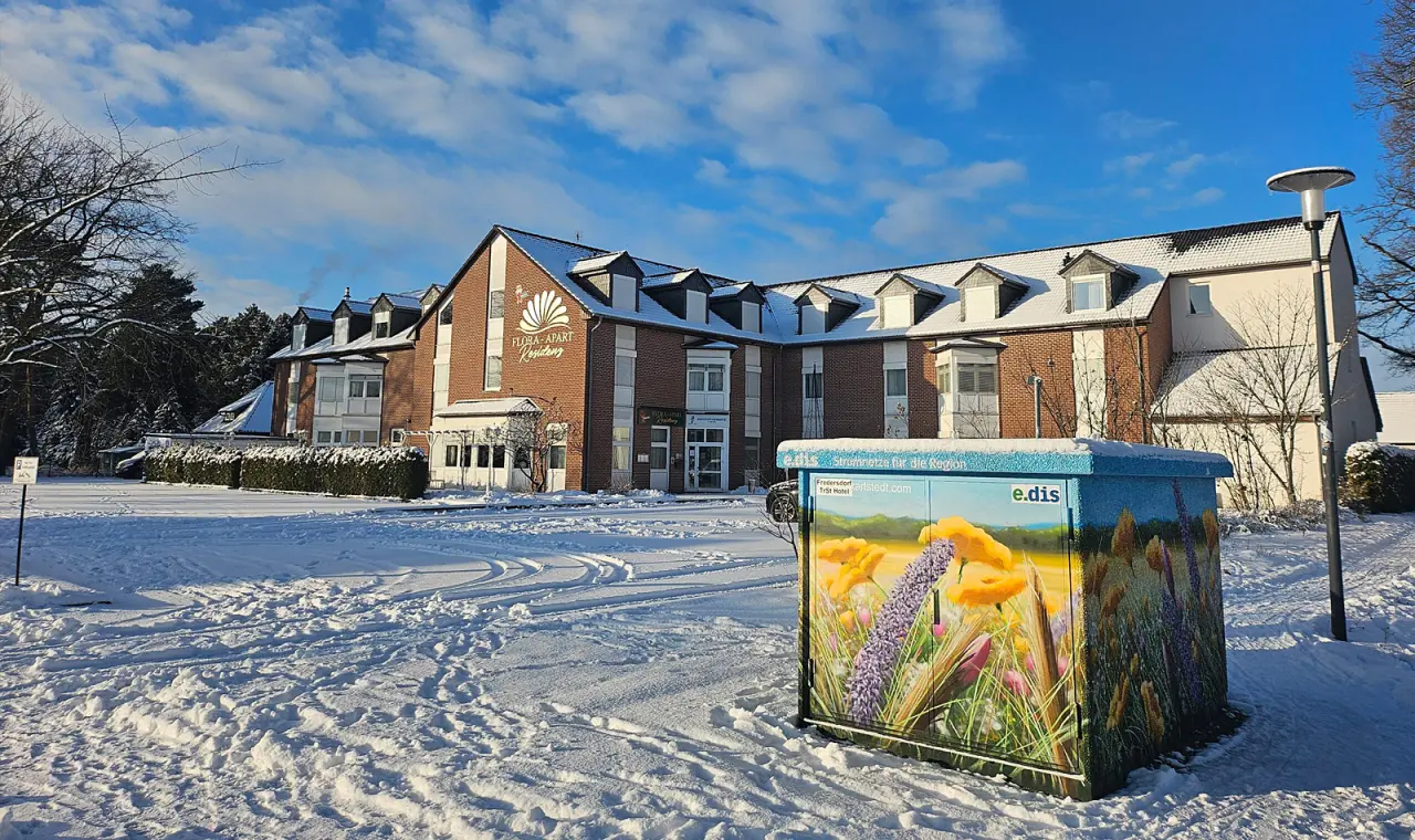 Aussenansicht des Hotel Flora-Apart Residenz im Winter mit verschneiter Zufahrt und blauem Himmel.