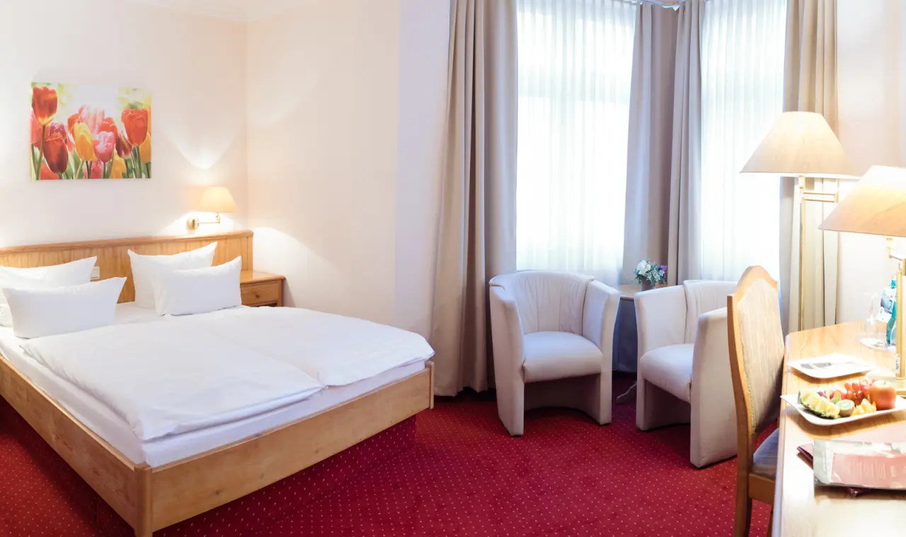 Gemütliches Doppelzimmer im Hotel Flora mit Holzbett, rotem Teppich und Sitzbereich am Fenster.