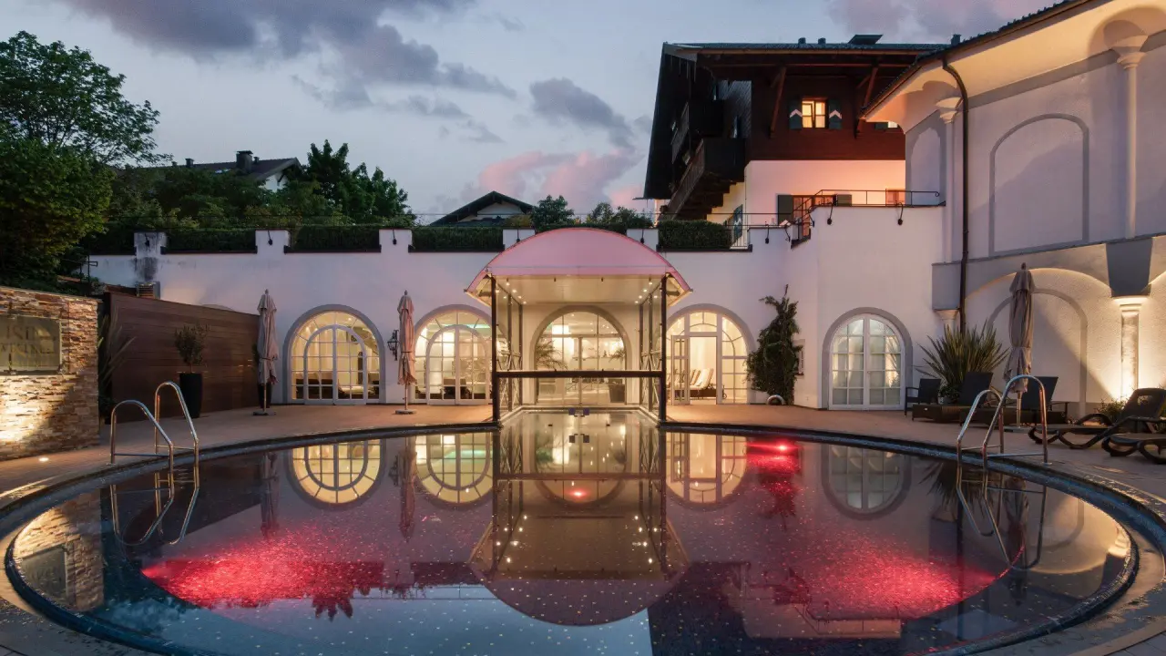 Beheizter Aussenpool mit roter Beleuchtung und Hotelgebäude bei Dämmerung im Wellnessbereich von Gut Ising.