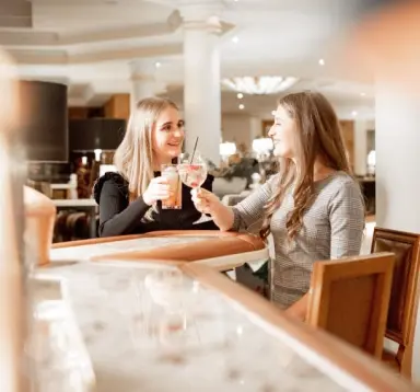 Zwei lachende Frauen stoßen mit Cocktails an der Bar des Avita Resort****Superior an, gemütliche Atmosphäre.