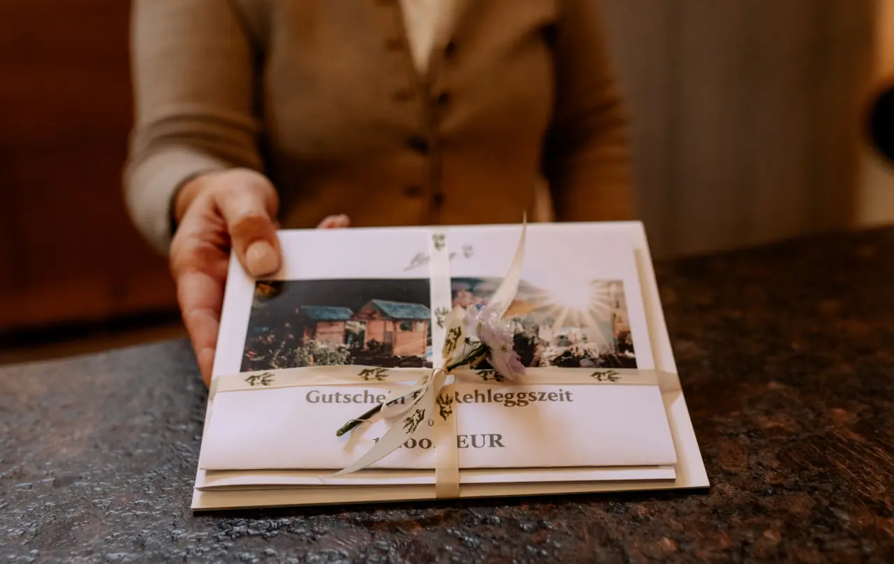 Geschenkgutschein für eine 'Rehleggszeit' im Berghotel Rehlegg, mit Band und Blüte verziert, von einer Hand präsentiert.