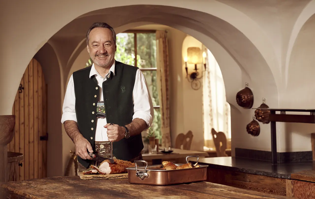 Küchenchef in Tracht tranchiert Braten in der rustikalen Küche des Berghotel Rehlegg.