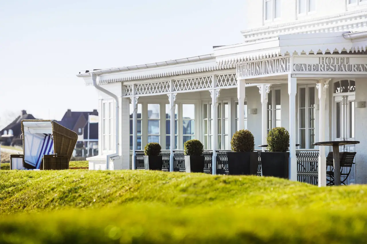 Weißes Fährhaus Sylt Hotelgebäude mit traditioneller Architektur, Veranda, grünem Rasen und Strandkorb unter blauem Himmel.