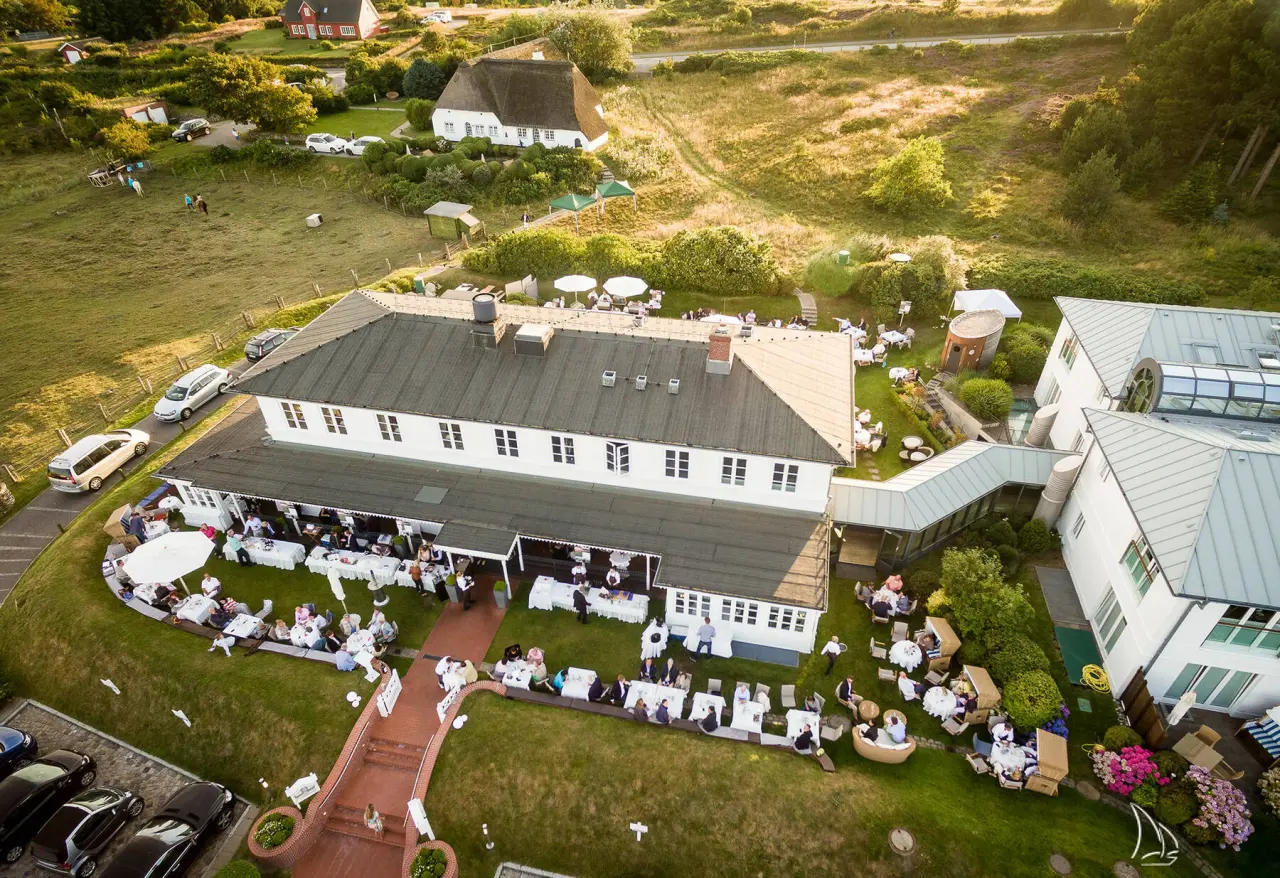 Luftaufnahme des Fährhaus Sylt mit Hotelgebäuden, weitläufigem Garten und Gästen bei einem Event im Freien.