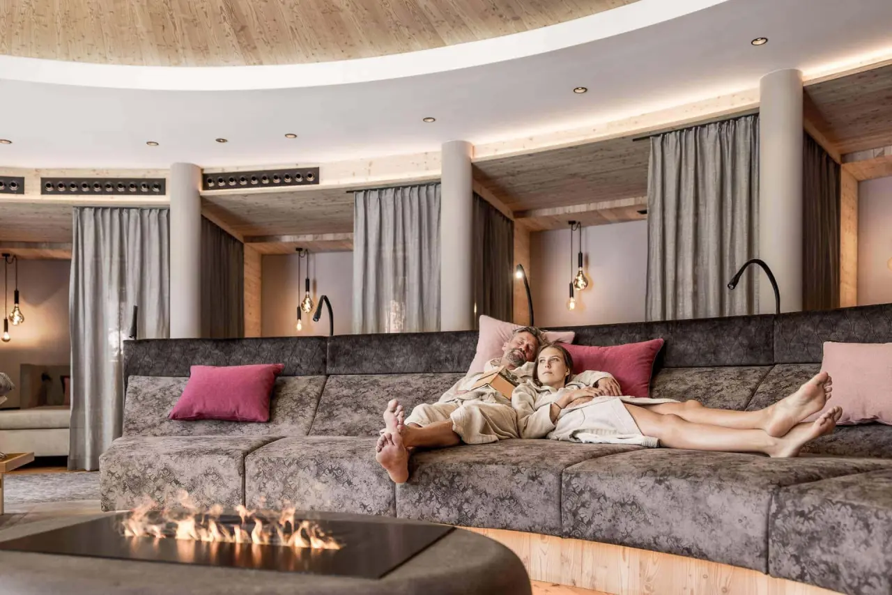 Entspannendes Paar in Bademänteln auf einer gemütlichen Lounge-Couch mit Kamin im Wellnessbereich des Holzleiten Bio Wellness Hotel.
