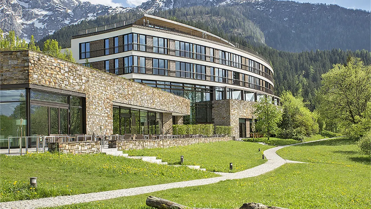 Modernes Kempinski Hotel Berchtesgaden mit Natursteinfassade und Bergpanorama, umgeben von gruenen Wiesen und Baeumen.