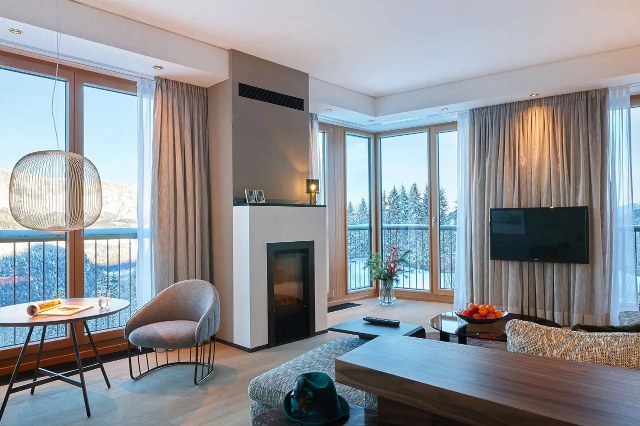 Luxuriöses Wohnzimmer einer Suite im Kempinski Hotel Berchtesgaden mit Kamin und Panoramablick auf die verschneite Berglandschaft.