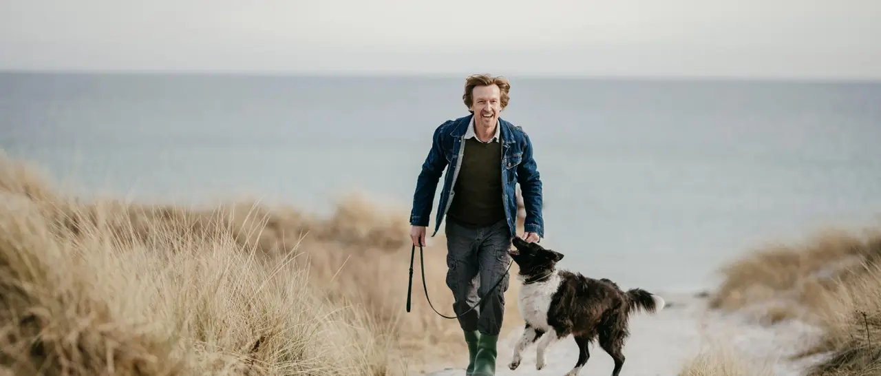 Lachender Mann mit Hund beim Spaziergang auf einem Dünenweg am Strand des Strandhotel Kurhaus Juist mit Meerblick.