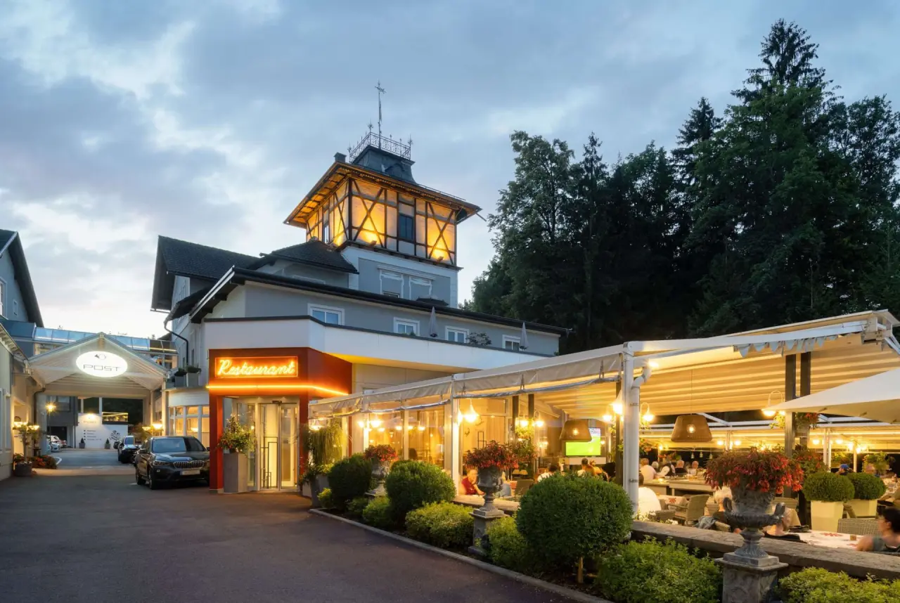 Abendliche Aussenansicht des Hotel Post Velden mit beleuchtetem Restaurant und Terrasse unter bewölktem Himmel.
