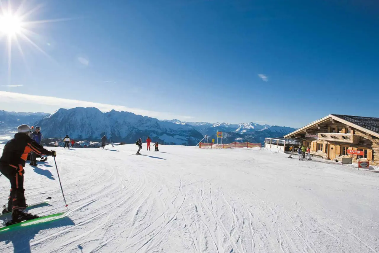 Sonniger Skitag auf der Piste mit Blick auf die verschneiten Alpen und eine urige Skihuette, ideal fuer Gaeste des Hotel DER HECHL.
