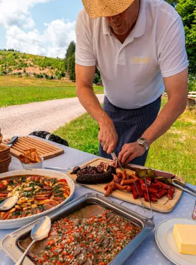 Mann schneidet Wurst und Fleisch an einem Buffet im Freien, mit weiteren Speisen und grüner Landschaft im Hintergrund, Hotel Kesslermühle