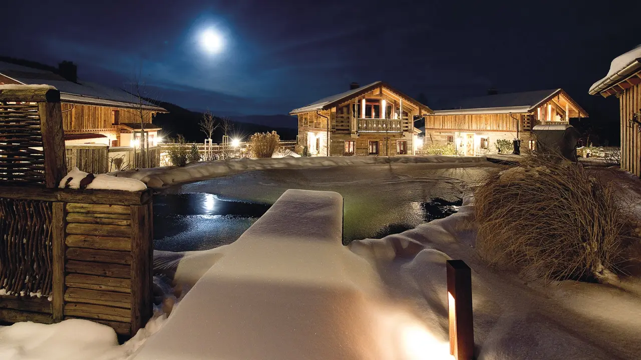 Winterliche Nachtansicht des Burghotel Sterr mit beleuchteten Holzgebäuden und einem schneebedeckten Aussenpool unter Vollmond.