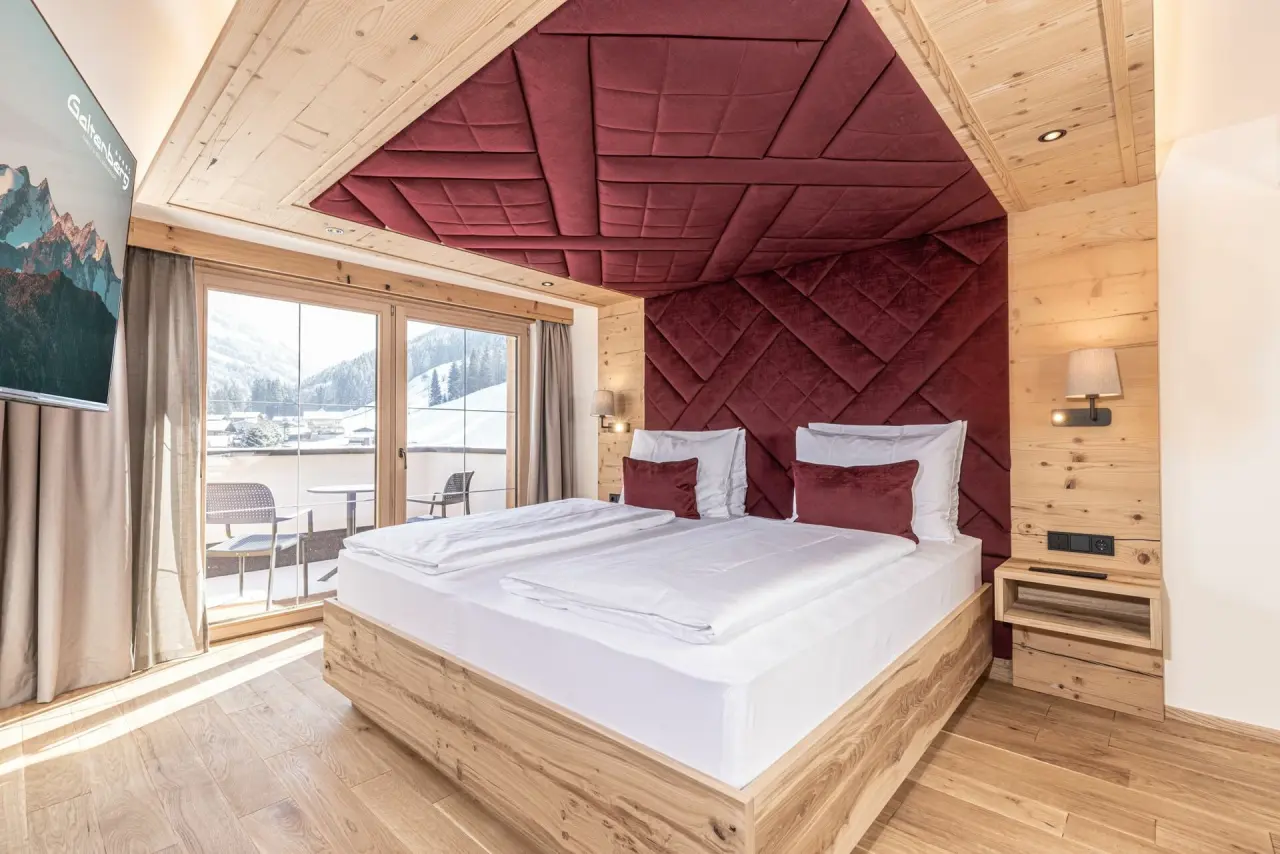 Modernes Doppelzimmer im alpinen Stil mit Holzelementen, rotem Kopfteil und Balkonblick im Galtenberg Resort 4*S.