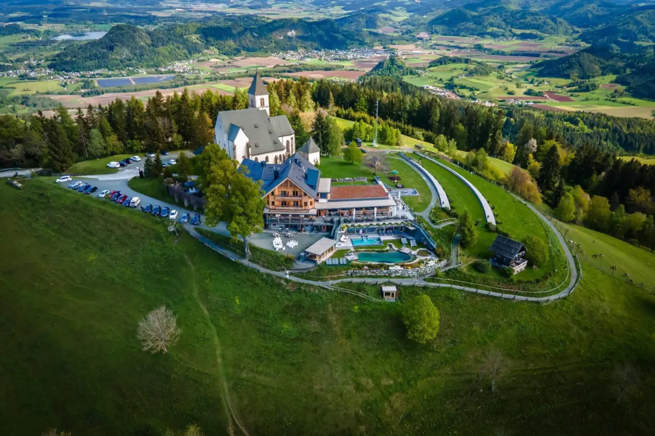 Gipfelhaus Magdalensberg: Panoramablick auf das Hotel, Kirche und Aussenpools inmitten der gruenen Berglandschaft.