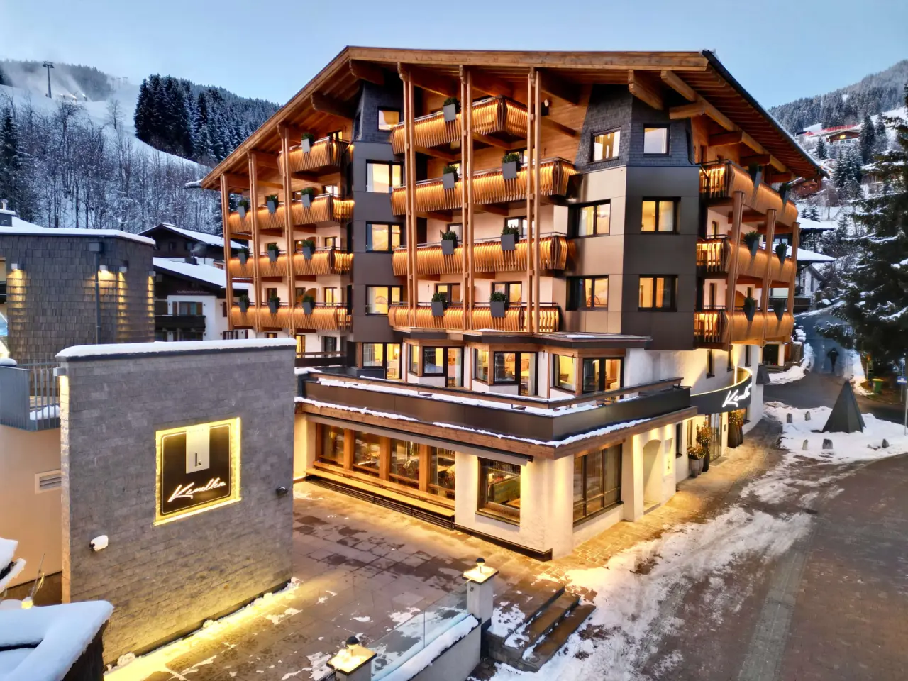 Beleuchtetes Hotel Kendler im Winter mit verschneiter Berglandschaft und Skipiste im Hintergrund.
