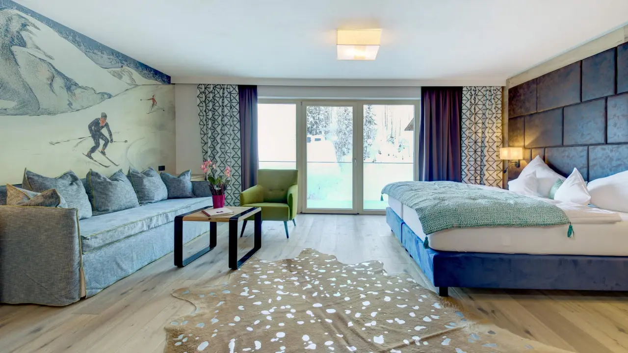 Gemütliches Hotelzimmer im Hotel Kendler mit Doppelbett, Sofa, Skifahrer-Wandbild und Balkonblick auf verschneite Bäume.
