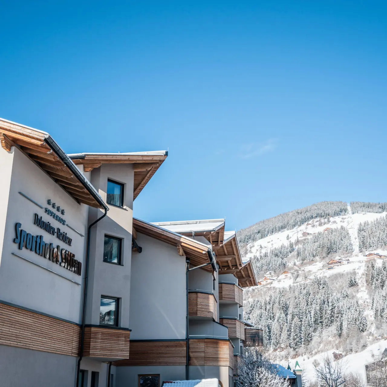 Aussenansicht des Dolomiten Residenz Sporthotel Sillian im Winter mit verschneiten Bergen und blauem Himmel.