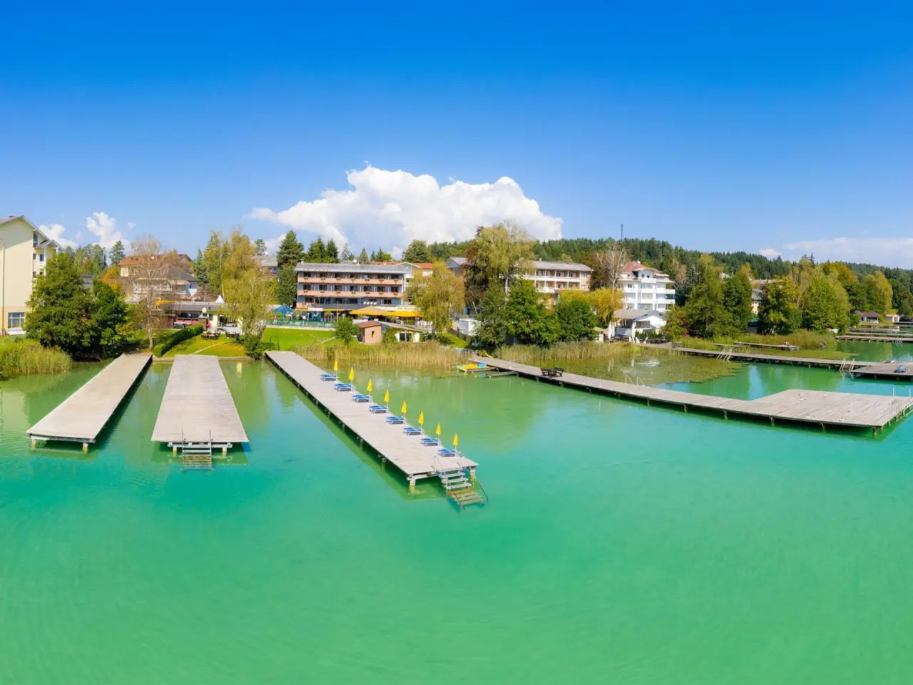 Luftaufnahme des Golf-Tennis-Wellnesshotel Mori direkt am türkisblauen See mit Badestegen und Liegen unter blauem Himmel.