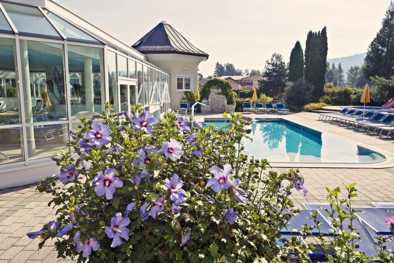 Aussenpoolbereich des Golf-Tennis-Wellnesshotel Mori mit blühenden Sträuchern, Liegestühlen und Glaspavillon.