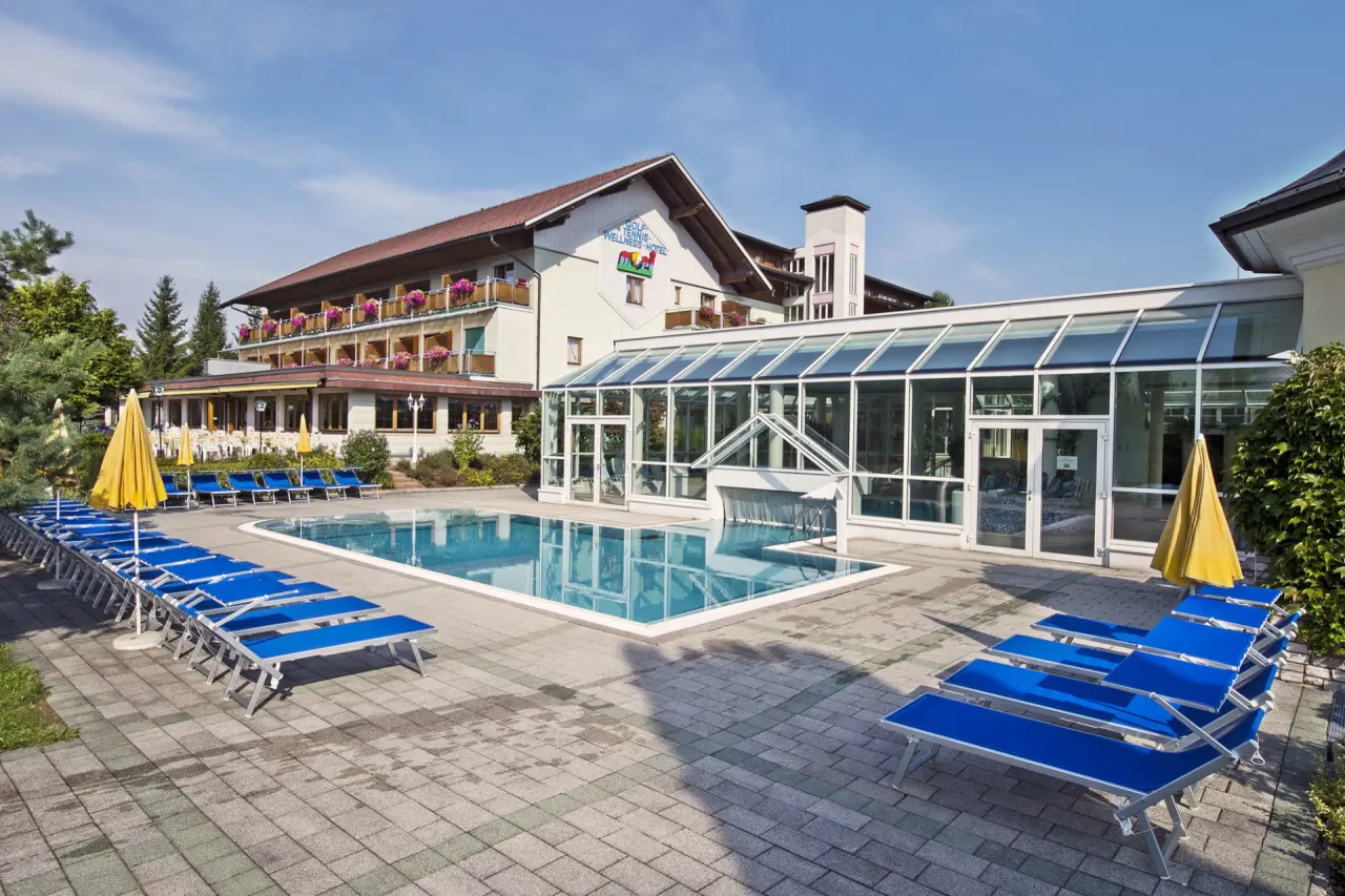 Sonniger Aussenpoolbereich mit blauen Liegen und dem Hauptgebäude des Golf-Tennis-Wellnesshotel Mori.