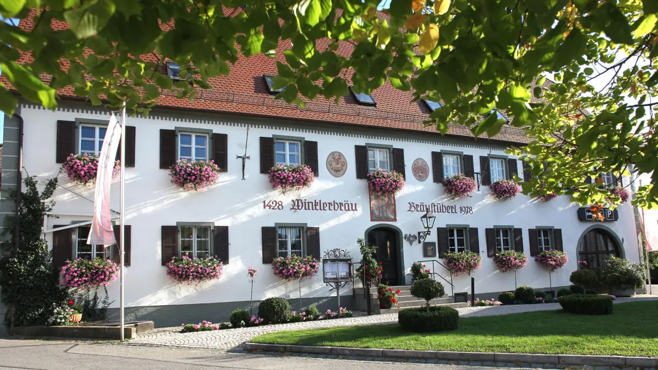 Historische Fassade des Winkler Bräu Gutshofhotel mit Blumenkästen und grünen Fensterläden in der Sonne.