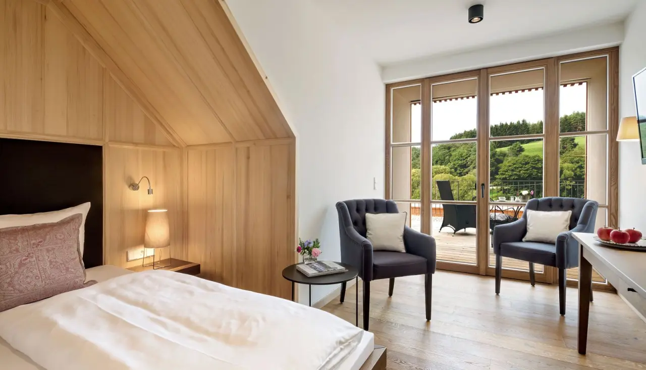 Gemütliches Hotelzimmer im Winkler Bräu Gutshofhotel mit Holzelementen, Sitzbereich und Balkon mit Blick ins Grüne.