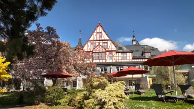 Historisches Moselschlösschen Spa & Resort mit blühendem Garten, roten Sonnenschirmen und Liegestühlen unter blauem Himmel.