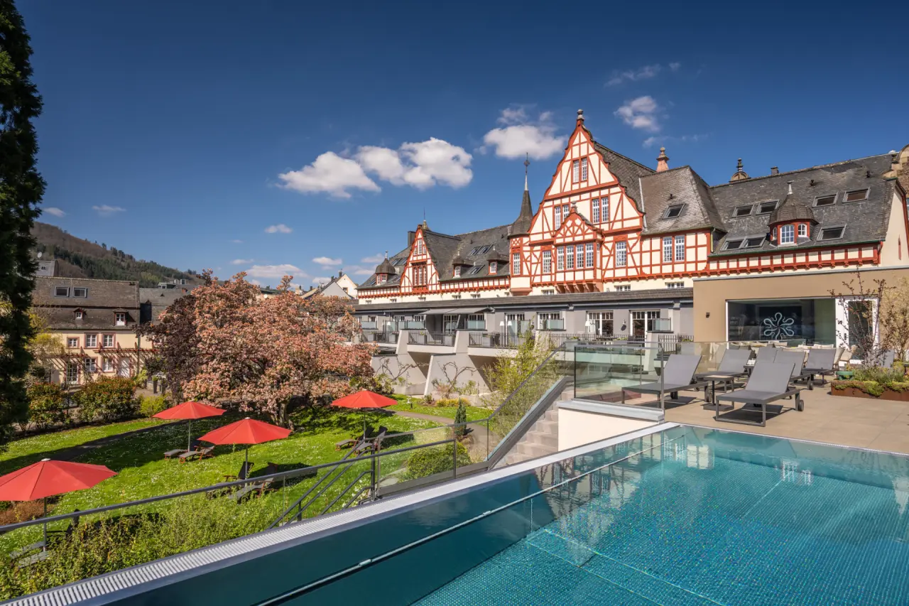 Aussenansicht des Moselschlösschen Spa & Resort mit historischem Fachwerk, modernem Aussenpool und blühendem Garten.