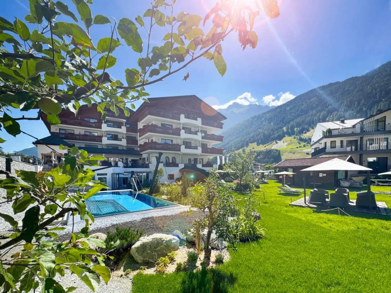 Aussenansicht des Hotel Der Stubaierhof mit Pool, Liegewiese und Alpenpanorama an einem sonnigen Tag.