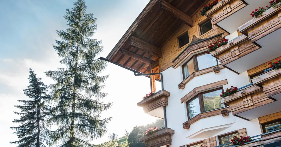 Fassade des Familien- & Gartenhotel Theresia****S mit Holzbalkonen, roten Blumen und hohen Tannen vor blauem Himmel.