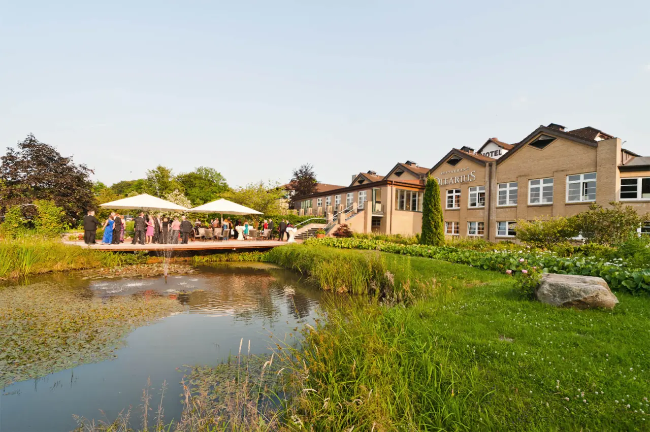 Aussenansicht des Hotel Waldschlösschen mit Teich, grüner Gartenanlage und einer Terrasse mit Gästen bei einer Veranstaltung.