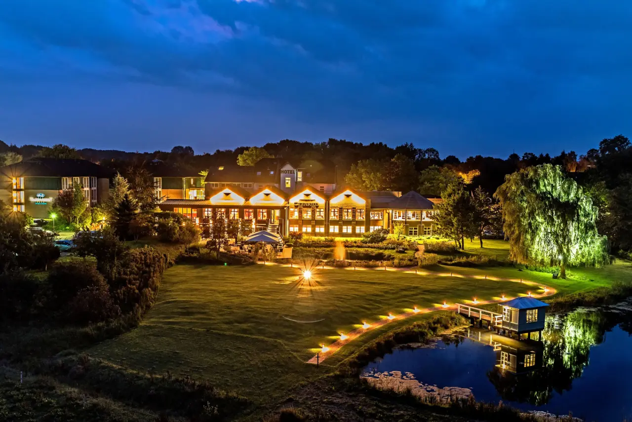 Nachtaufnahme des Hotel Waldschlösschen mit beleuchtetem Gebäude, Garten, Teich und Bootssteg unter blauem Himmel.