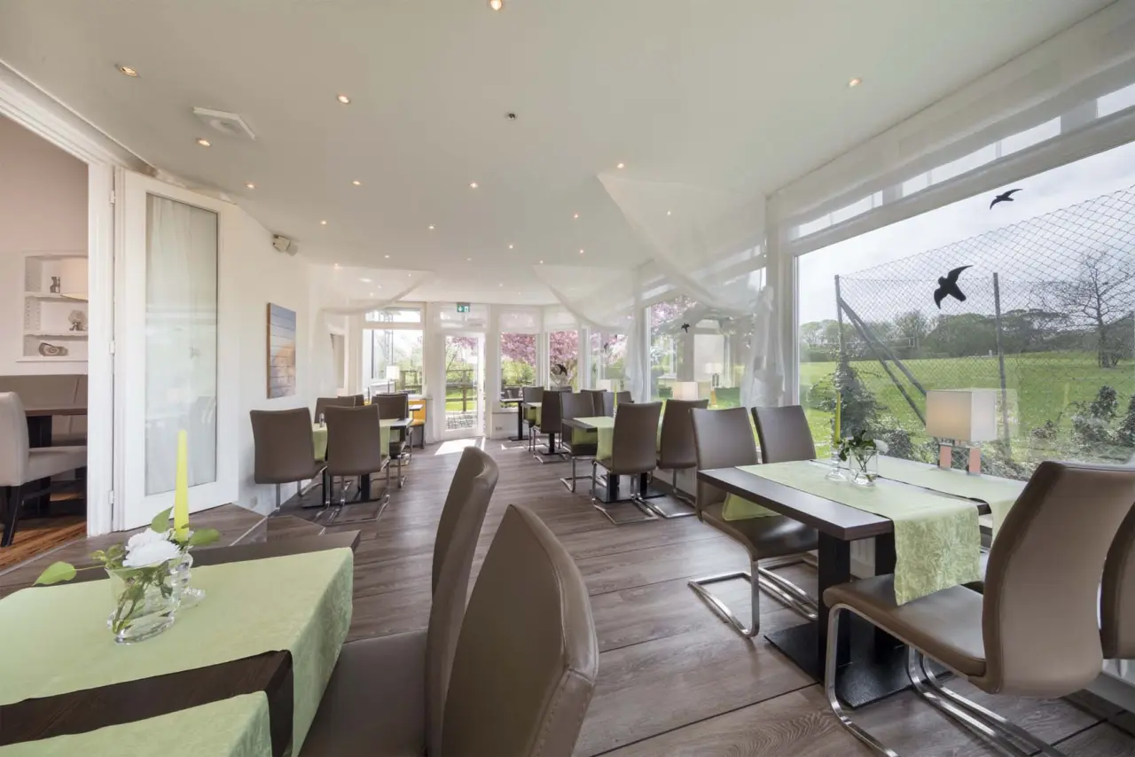 Heller Restaurantbereich mit großen Fenstern und Blick ins Grüne im HofHotel Krähenberg. Grüne Tischläufer und moderne Stühle.
