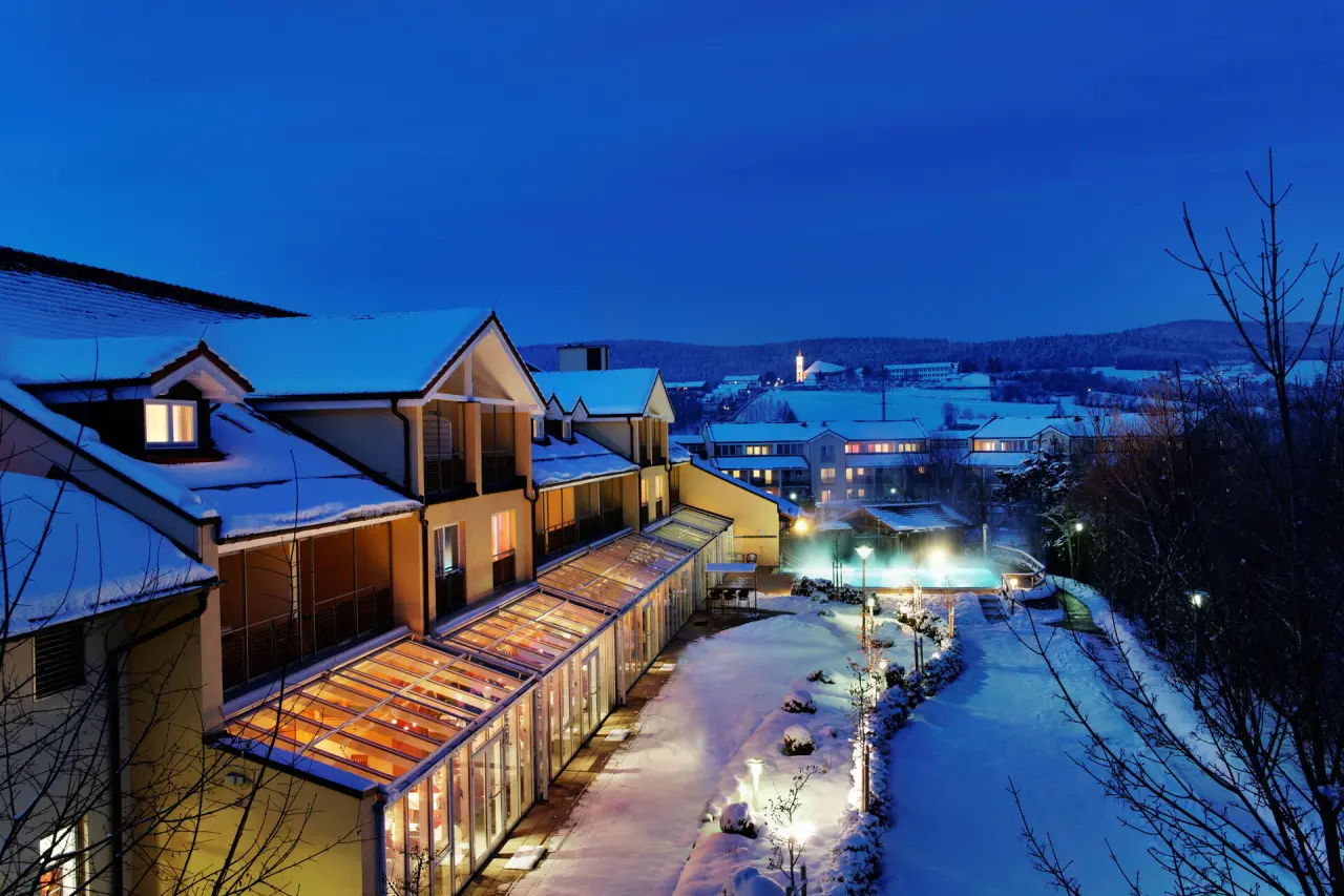 Winterliche Aussenansicht des Hotel Herzog Heinrich bei Dämmerung mit beleuchtetem Aussenpool und verschneiter Landschaft.