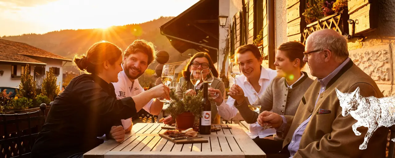 Fröhliche Gäste stoßen mit Wein auf der Restaurant-Terrasse des Romantik Hotel Spielweg bei Sonnenuntergang an.