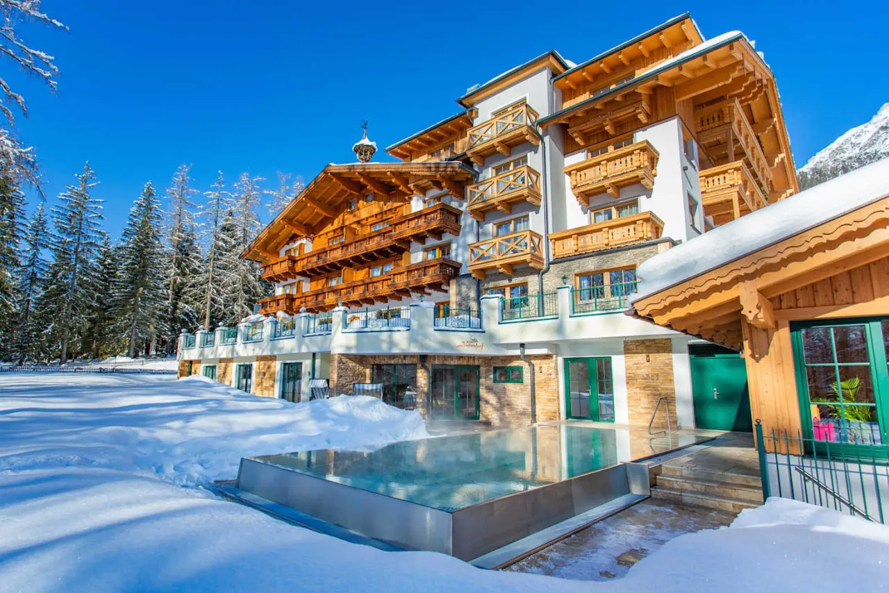 Alpines Hotel Lindenhof im Winter mit beheiztem Aussenpool, umgeben von verschneiter Landschaft und Tannen unter blauem Himmel.