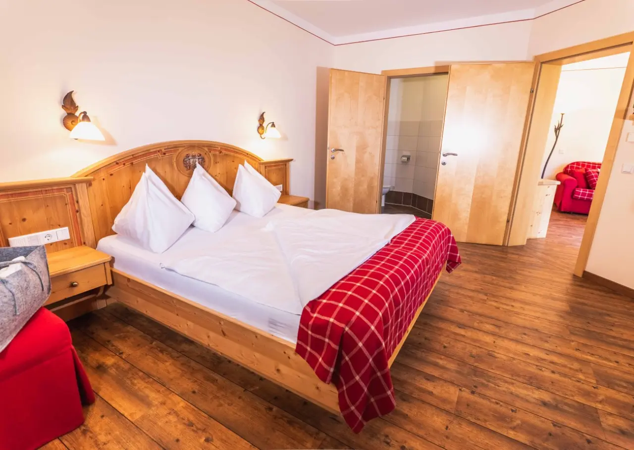 Gemütliches Doppelzimmer im Hotel Lindenhof mit Holzbett, roter Decke und Blick ins Bad und Wohnbereich.