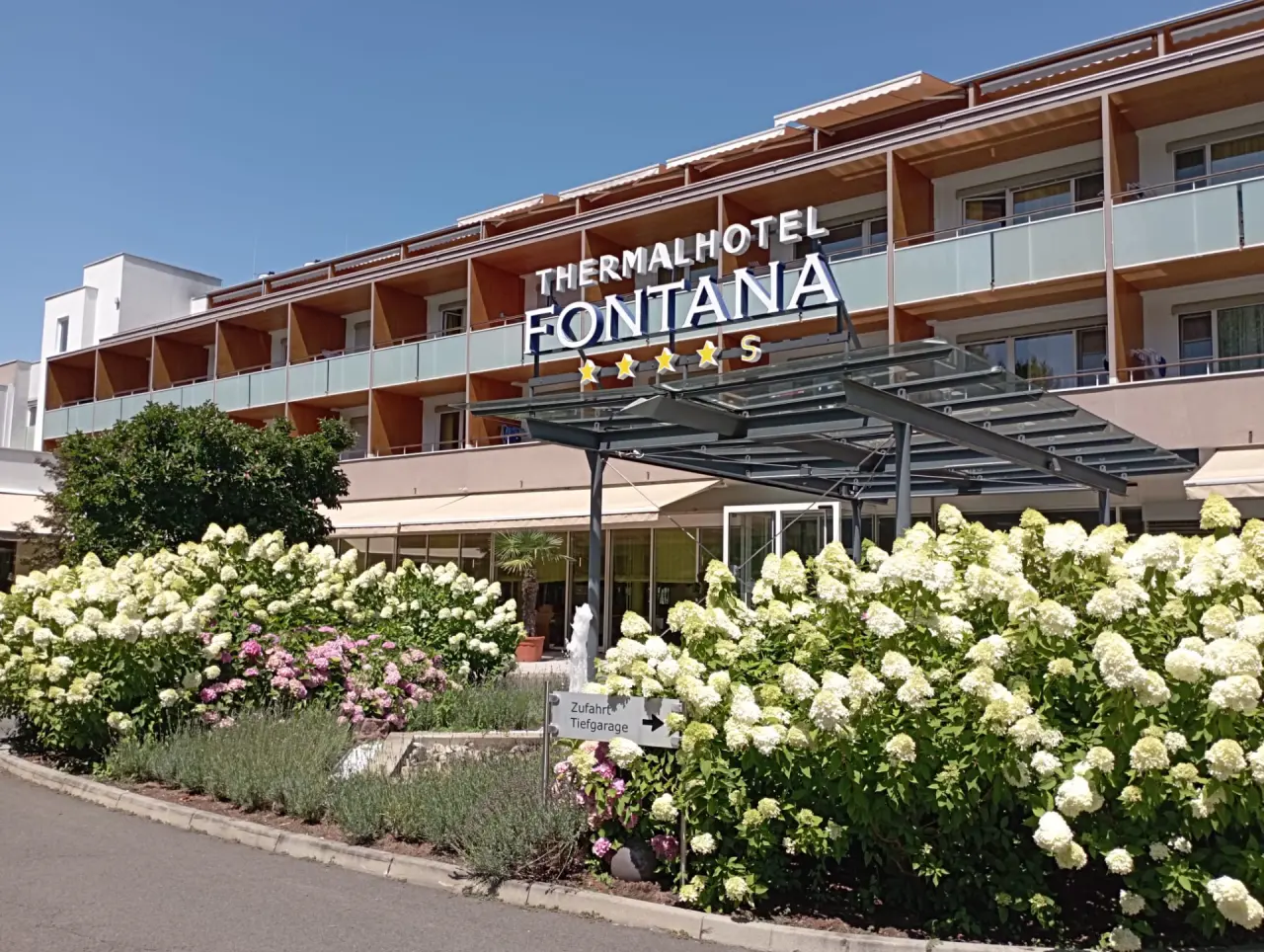 Aussenansicht des Thermalhotel Fontana mit blühenden Hortensien am Eingang und klarem blauen Himmel.