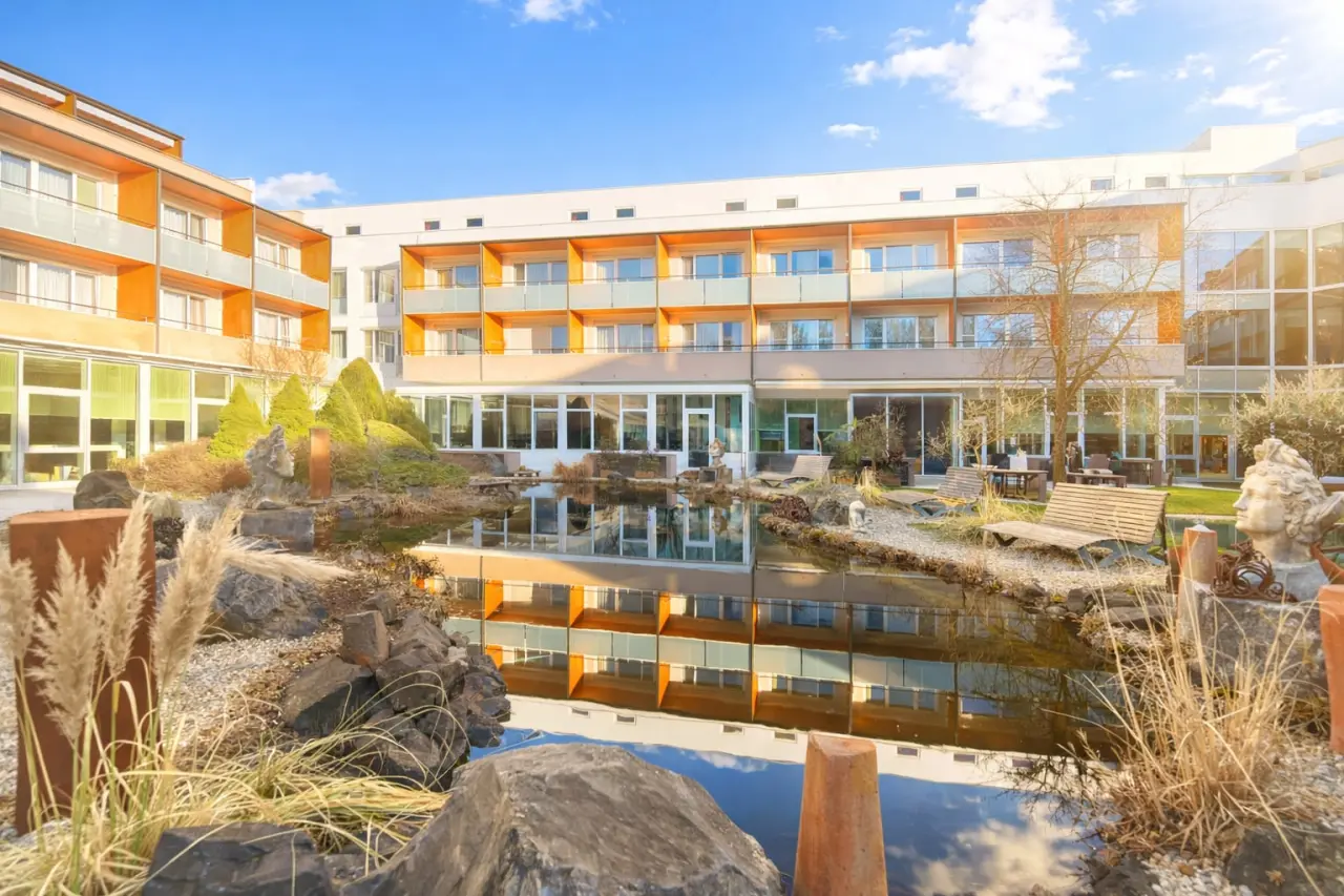 Aussenansicht des Thermalhotel Fontana mit einem idyllischen Teich im Garten, umgeben von Gruenflaechen und Sitzgelegenheiten unter blauem Himmel.