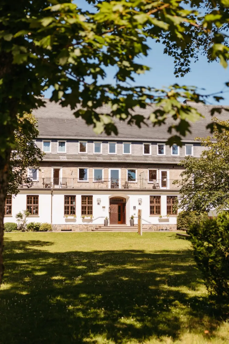 Aussenansicht des Berghotels Hoher Knochen im Sommer, umrahmt von gruenen Blaettern, mit weisser Fassade und Balkonen.